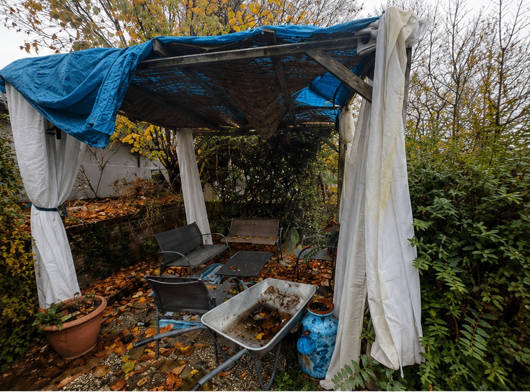 Avant : tonnelle bois vétuste recouverte d'une bâche bleue déchirée, sol en gravier jonché de feuilles mortes